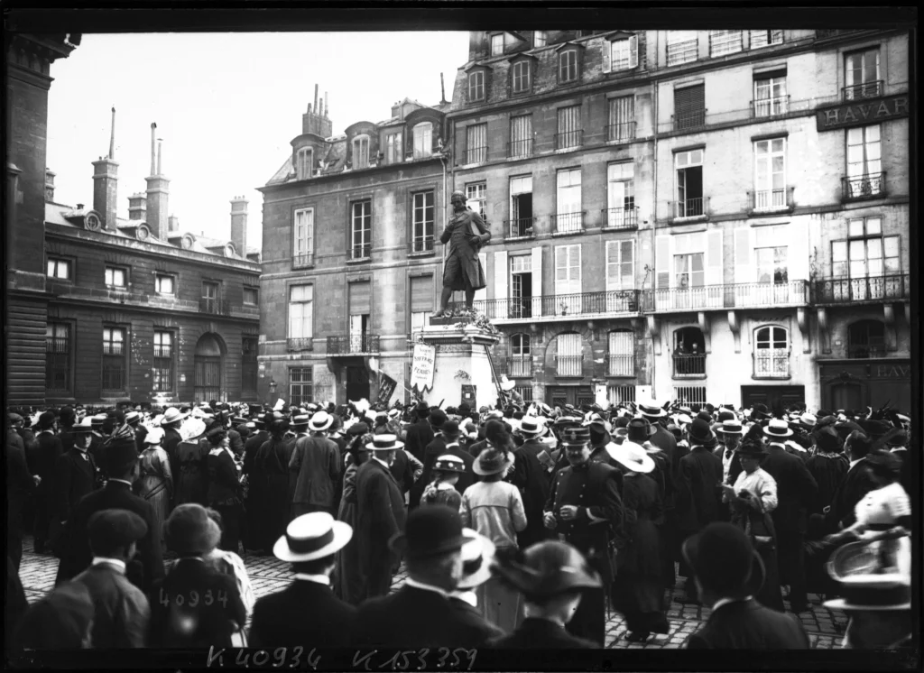 Photographie ancienne en noir et blanc. Une foule dense, coiffée de chapeaux, entoure la statue d'un homme en costume du XVIIIe siècle, sur un socle ; une pancarte est accrochée au socle. Des immeubles parisiens ferment la place ; un agent de police se tient dans la foule.